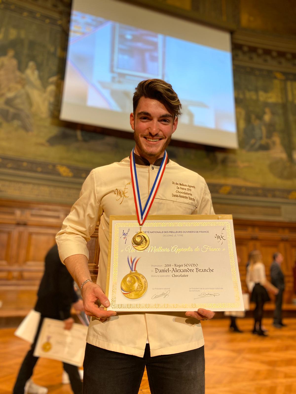 Photo de notre chef chocolatier quand il a reçu le prix du "meilleur apprenti de France 2019". Il tient à la main son diplôme et autour du coup la médaille d'or du concours.